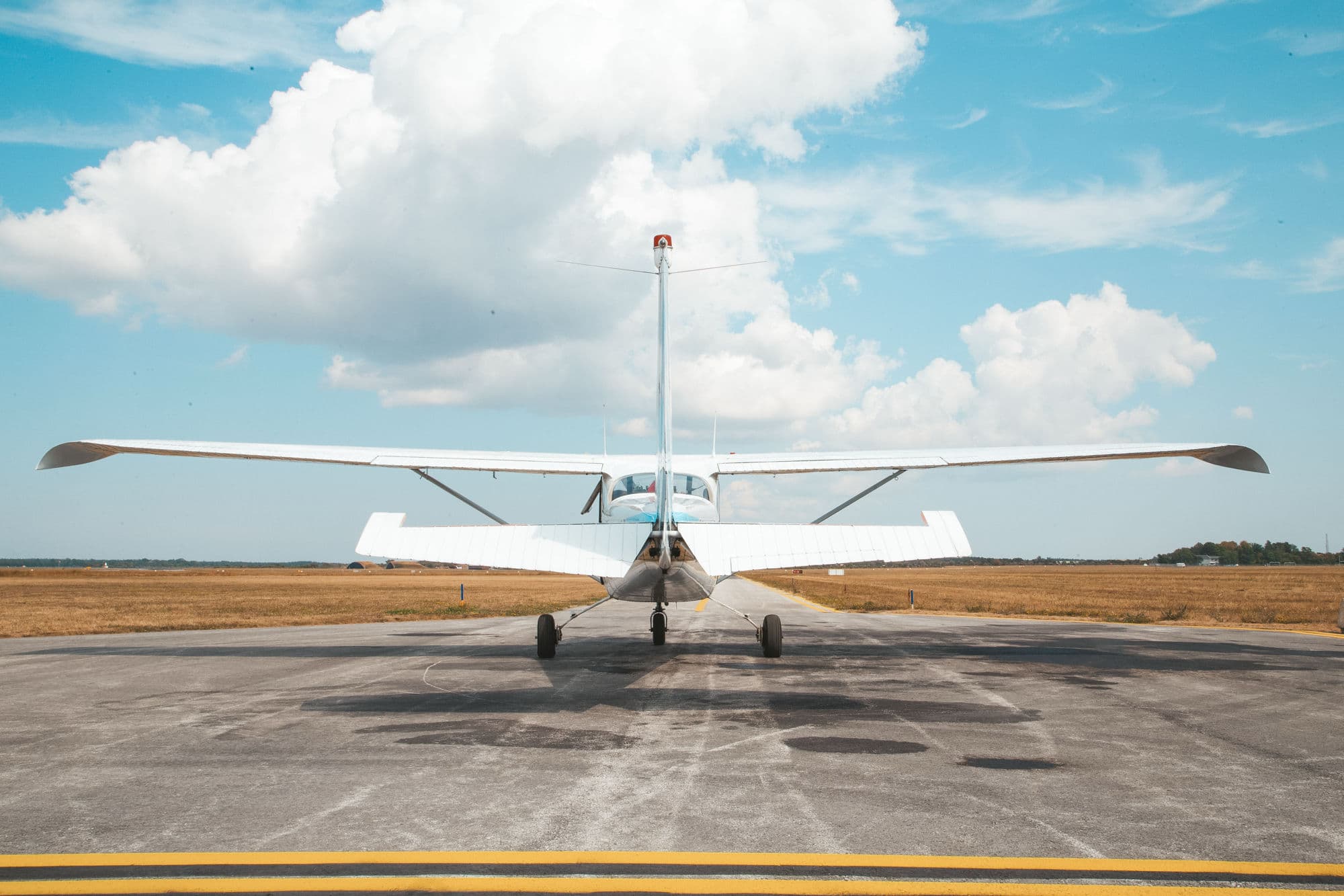 Avioneta branca de asa alta vista de trás na plataforma de um aeródromo, sob um céu de grandes cúmulos.